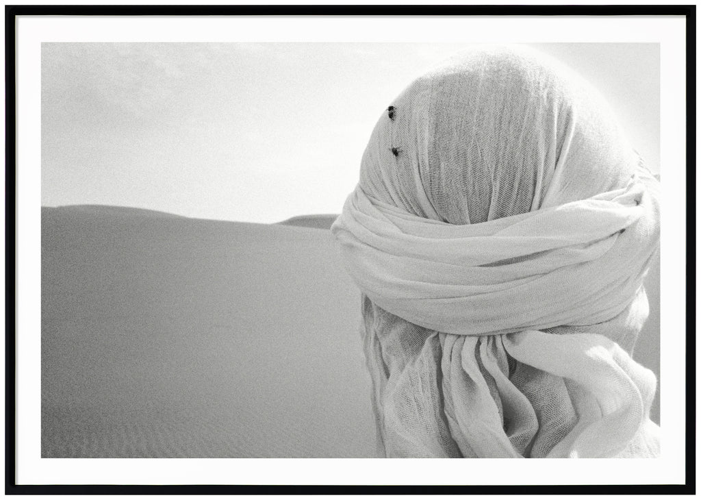 Black and white poster of close-up on head from behind with white shawl and two flies on in the desert. Black frame.
