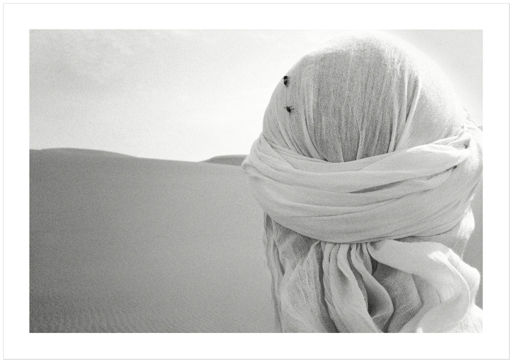 Black and white poster of close-up on head from behind with white shawl and two flies on in the desert.