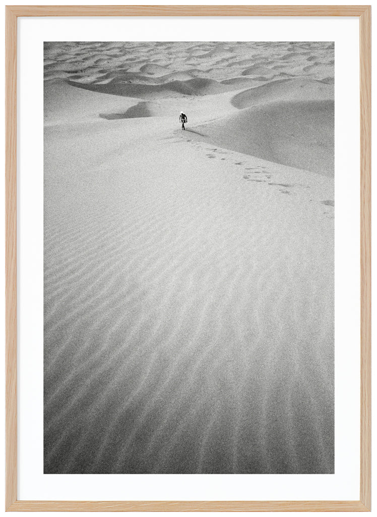 Black and white analog photograph of a man in the distance, walking up a huge sand dune in the middle of the Sahara desert. Oak frame. 