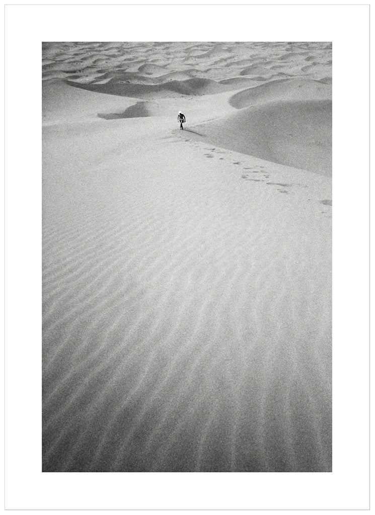 Black and white analog photograph of a man in the distance, walking up a huge sand dune in the middle of the Sahara desert.
