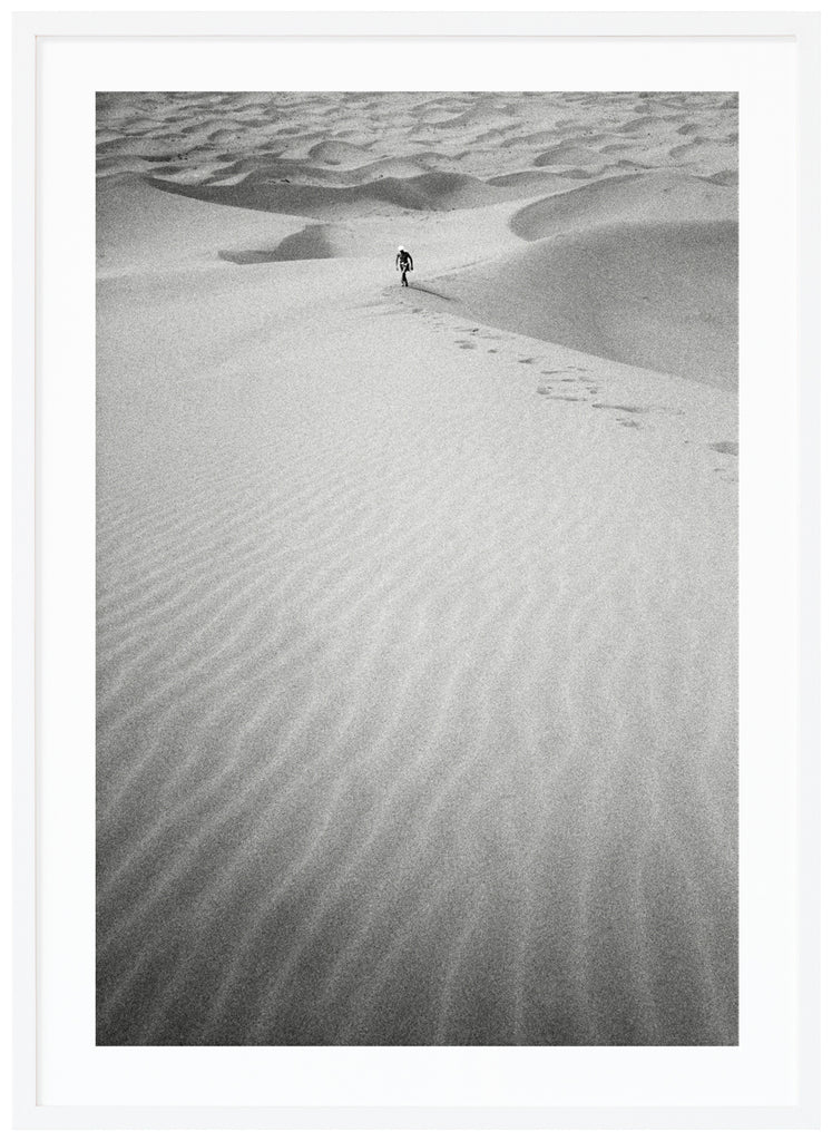 Black and white analog photograph of a man in the distance, walking up a huge sand dune in the middle of the Sahara desert. White frame. 