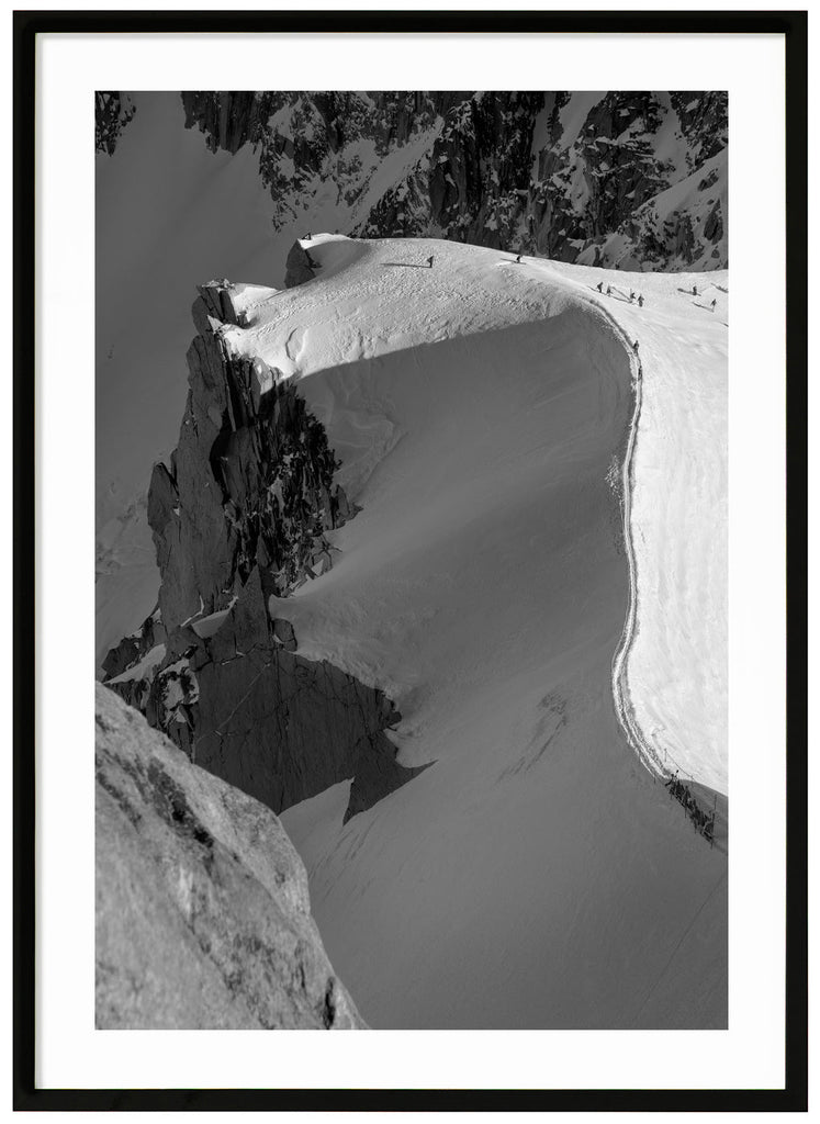 Black and white photograph of skiers hiking on snow-capped mountain ridge in the French Alps.  Black frame. 