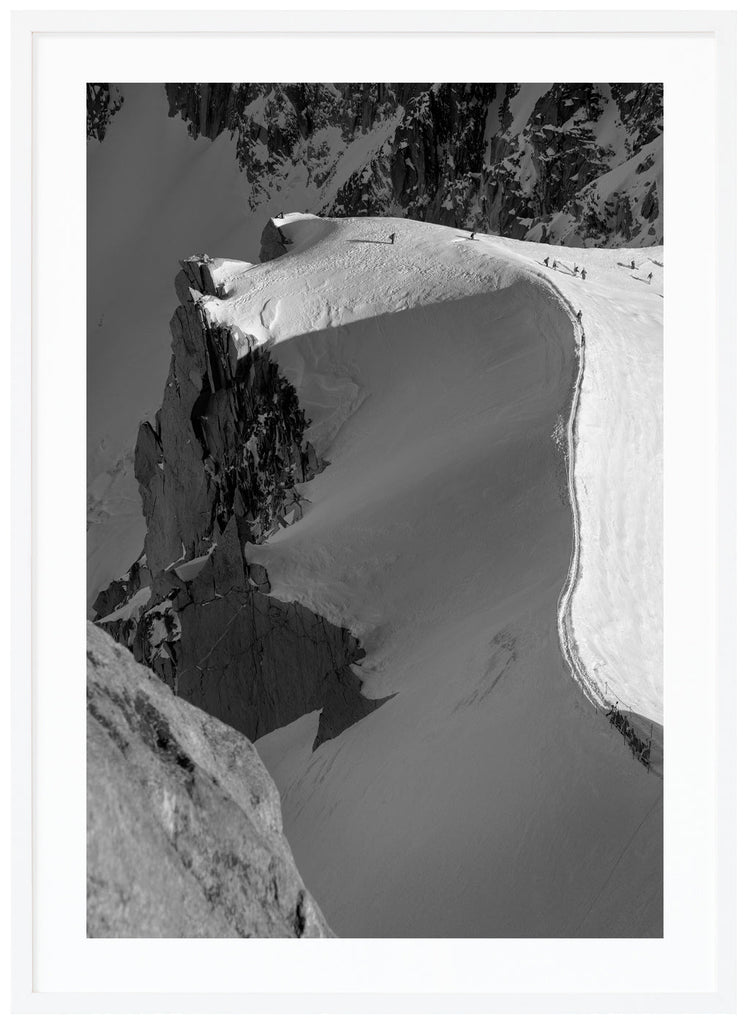 Black and white photograph of skiers hiking on snow-capped mountain ridge in the French Alps. White frame. 